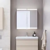 Liten baderomsinspirasjon med servantskap og overskap over toalett i beige spesialfarge, elegant speilskap med integrert lys, de lyse fargene og hvite gardinet skaper en romantisk og feminin atmosfære.