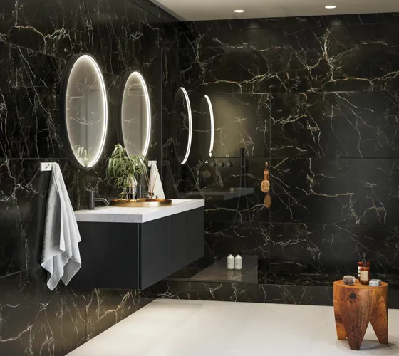 Elegant og moderne baderomsinspirasjon med mørke marmorerte vegger, runde opplyste speil med svart ramme, vegghengt servantskap med dobbeltvask og en romslig dusjniche