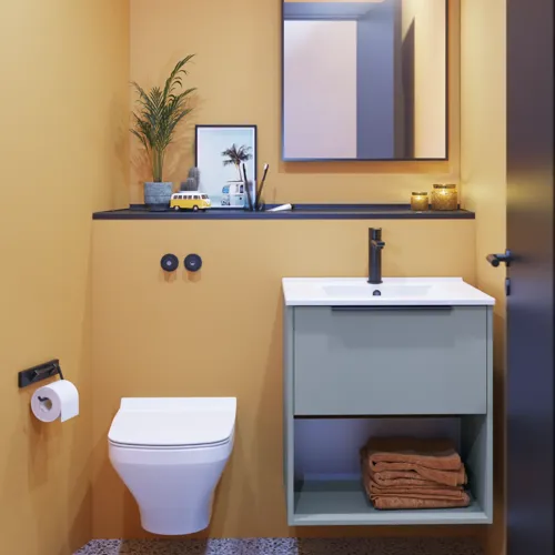 Liten baderomsinspirasjon med skrå og varme gule vegger, elegant vaskeskap i lys blå, tynn og stilren vask samt minimalistisk firkantet speil med svart kant og praktisk lampe.