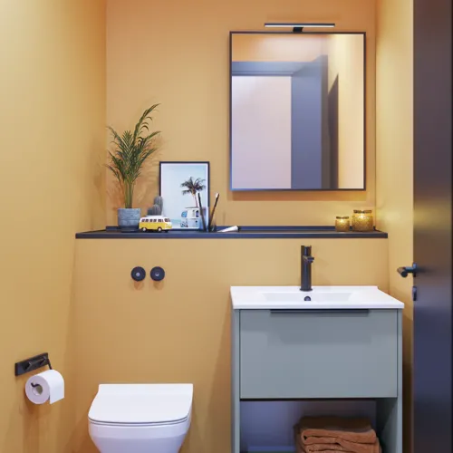 Liten baderomsinspirasjon med skrå og varme gule vegger, elegant vaskeskap i lys blå, tynn og stilren vask samt minimalistisk firkantet speil med svart kant og praktisk lampe.